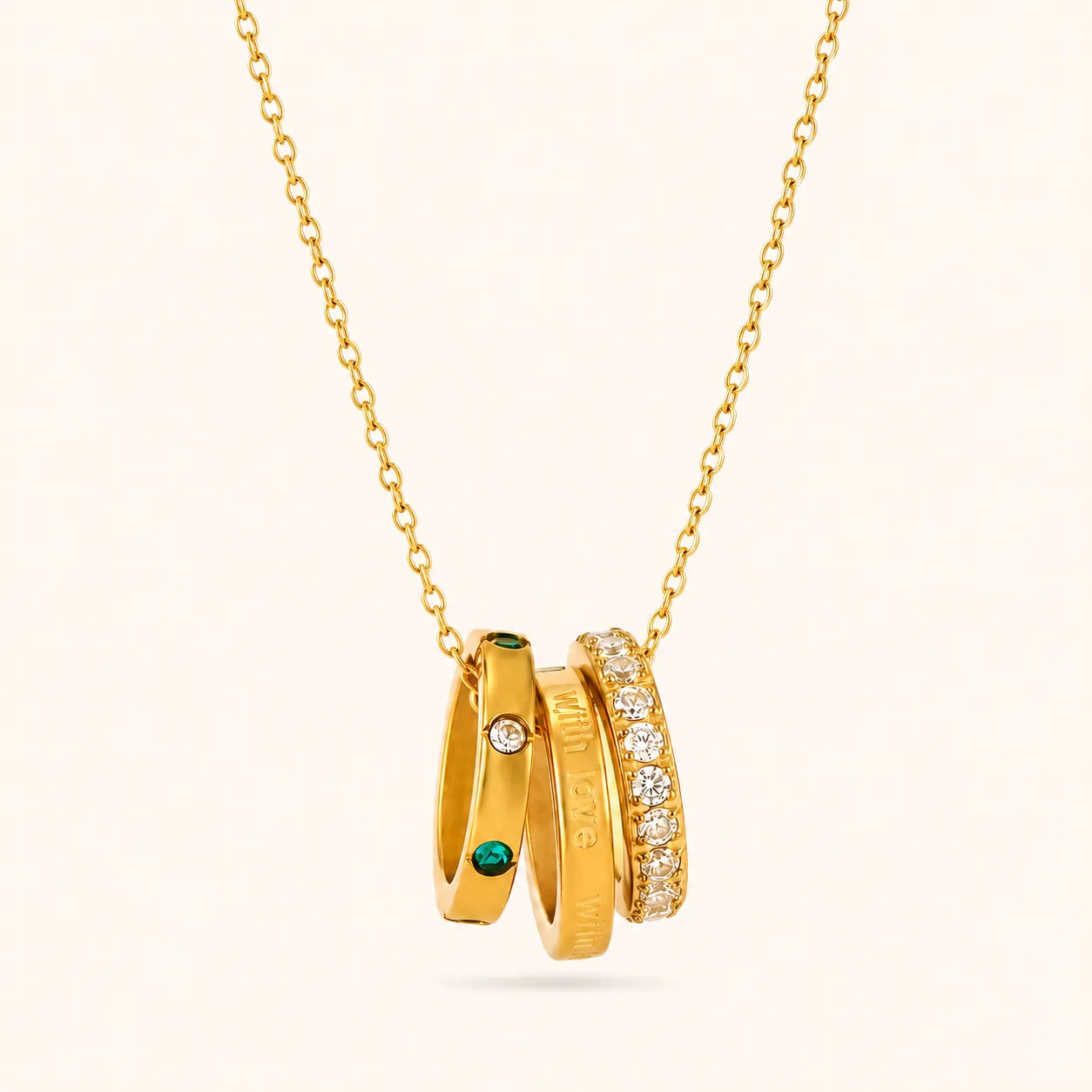 A gold chain necklace with a pendant featuring three interlocking gold rings, one adorned with a green emerald, one with a single clear stone, and one with a row of clear pavé stones, set against a light cream background.